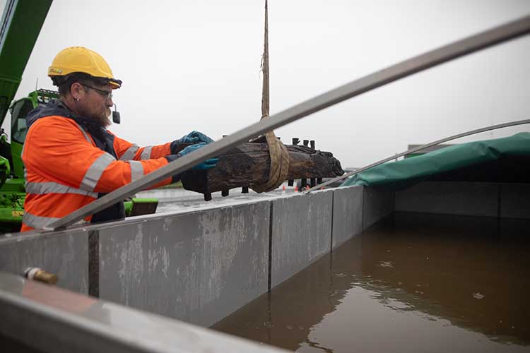 A person in hi-vis and a hard hat guides a crane carrying a timber plank into a steel tank of water