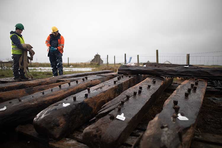 Two people wearing hi-vis and hard hats stand behind timber planks from the Sanday shipwreck 