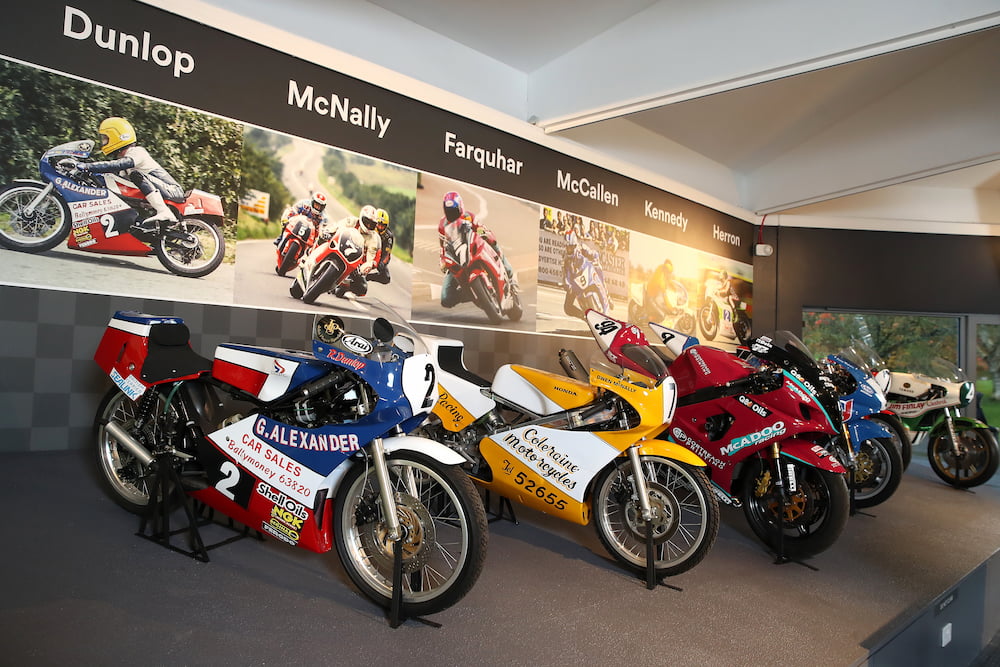 Five road racing motorbikes are on display in a line. On the wall behind them is the surname and photo of the person that road each bike 