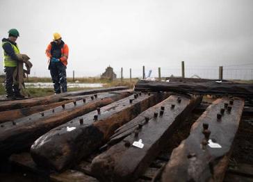 Two people wearing hi-vis and hard hats stand behind timber planks from the Sanday shipwreck 