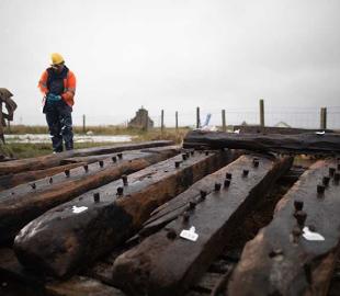 Two people wearing hi-vis and hard hats stand behind timber planks from the Sanday shipwreck 